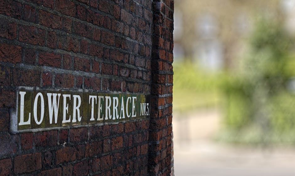 A close-up view of a brick wall displaying a metal street sign that reads 'LOWER TERRACE NW3' in white lettering on a green background. The brickwork is dark red with visible mortar joints, and the si